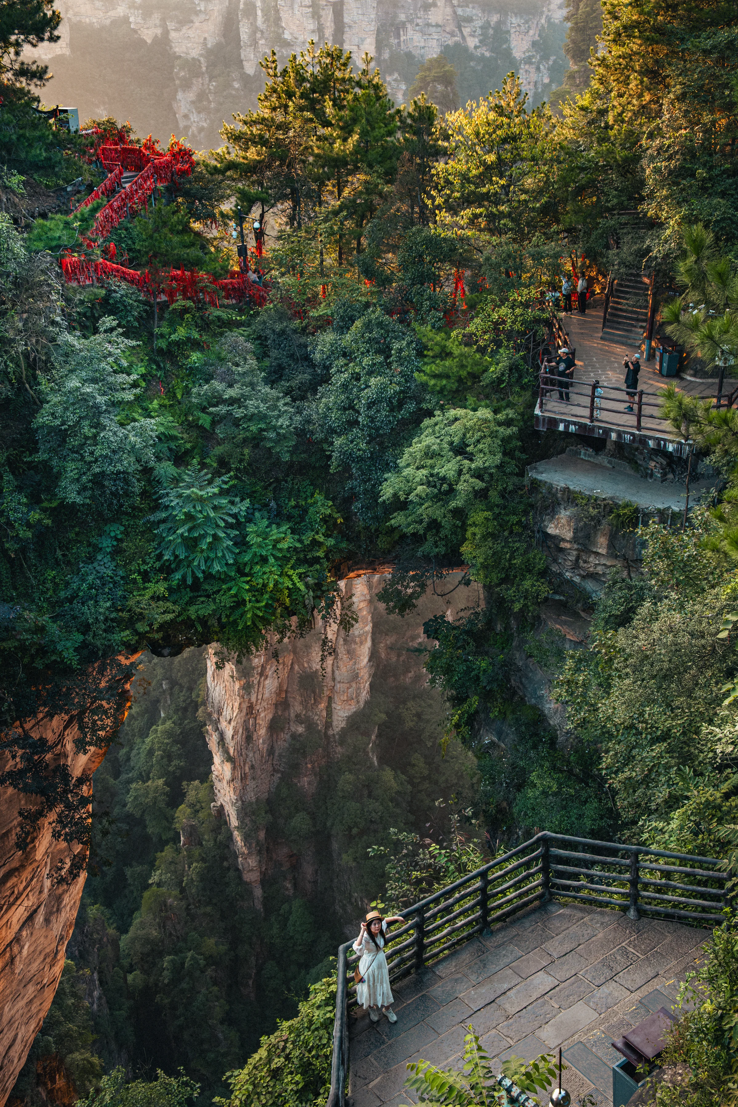 Woman in a white dress at the edge of a karst cliff, red prayer ribbons hanging in the upper left, Zhangjiajie landscape.