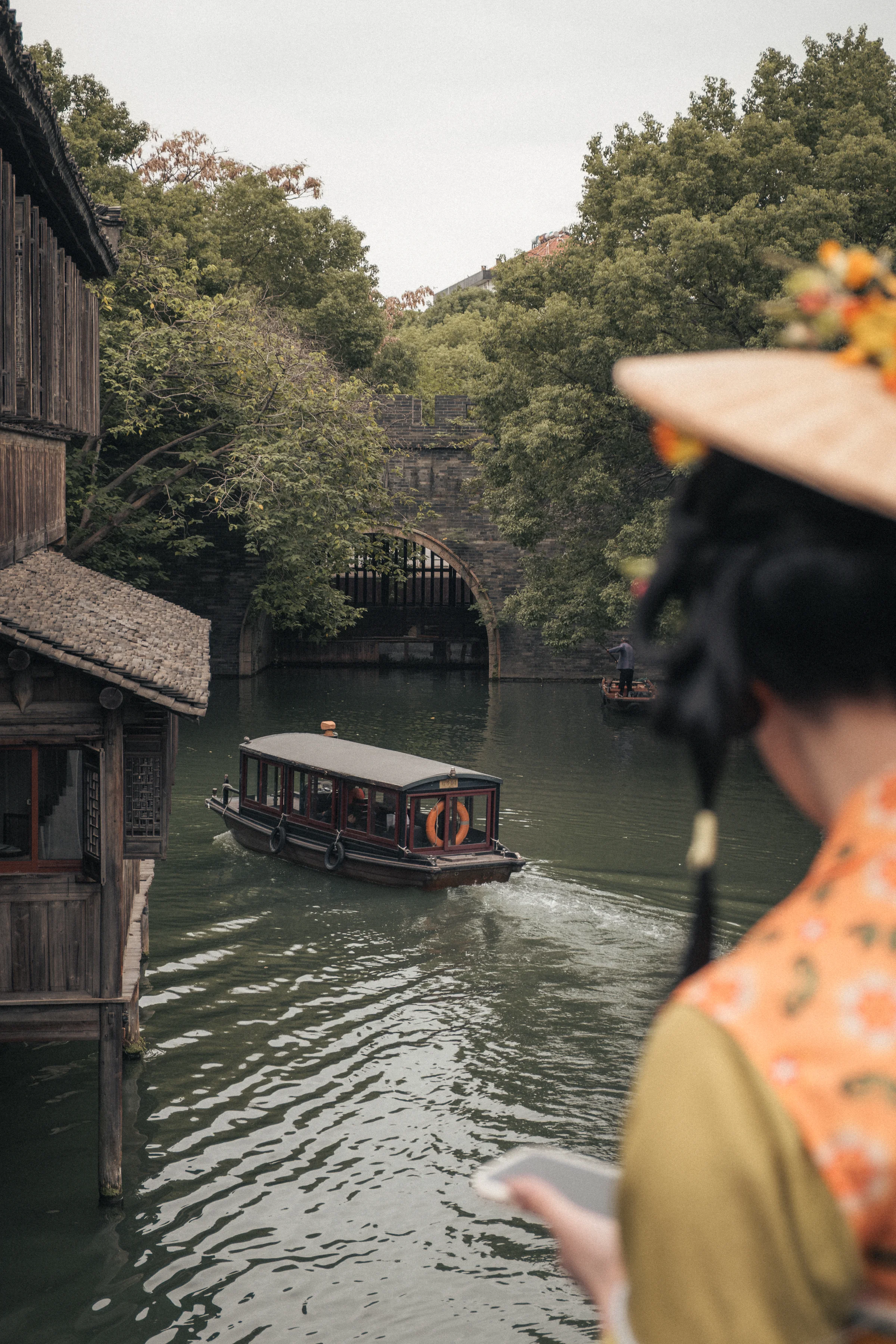 Young woman from behind wearing an orange hanfu and a yellow flowered hat, phone in hand, Wuzhen canal and traditional boat in the background.