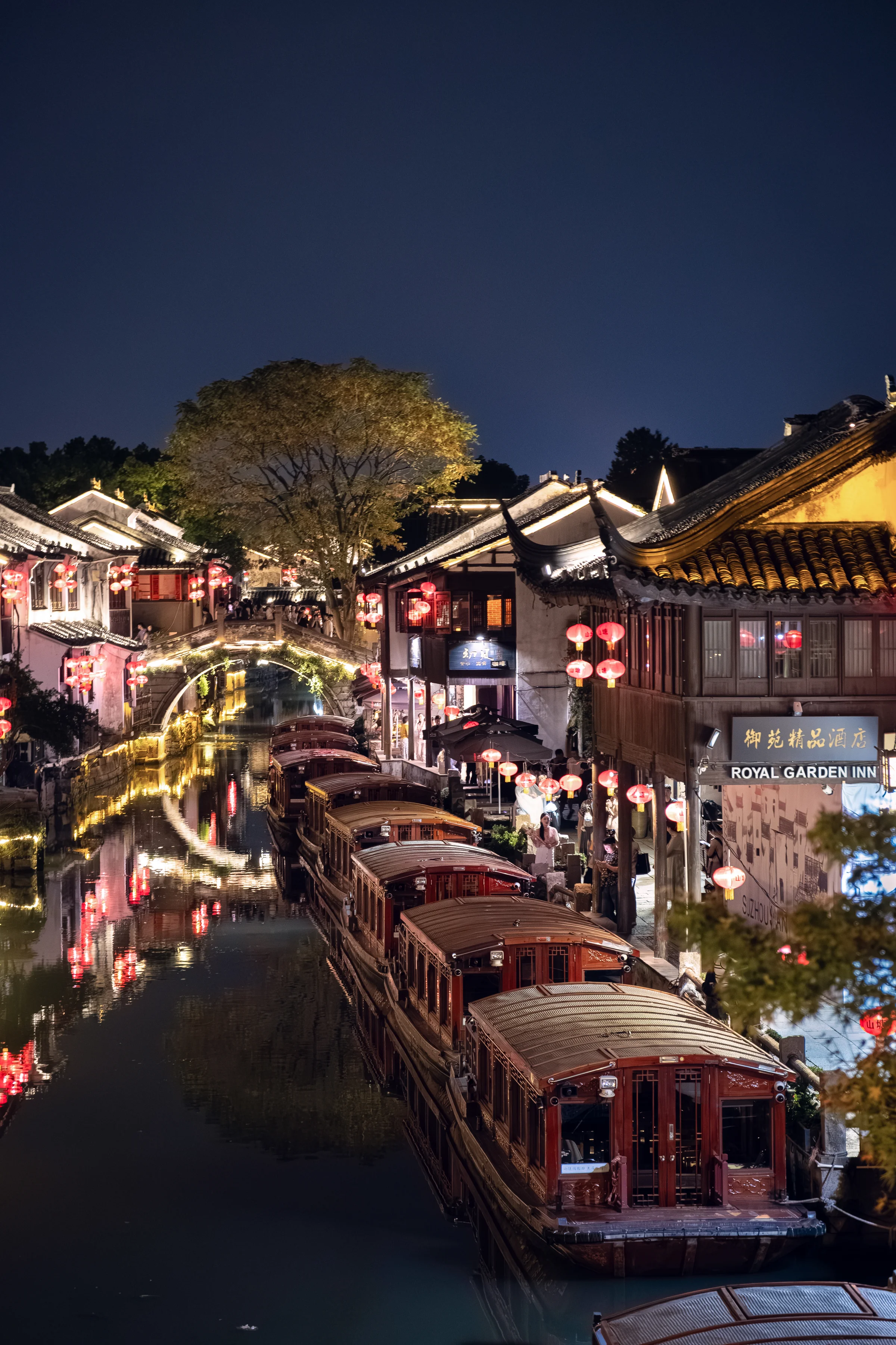 Suzhou canal at night, wooden boats lined up, red lanterns on the facades, lit arched bridge in the background.