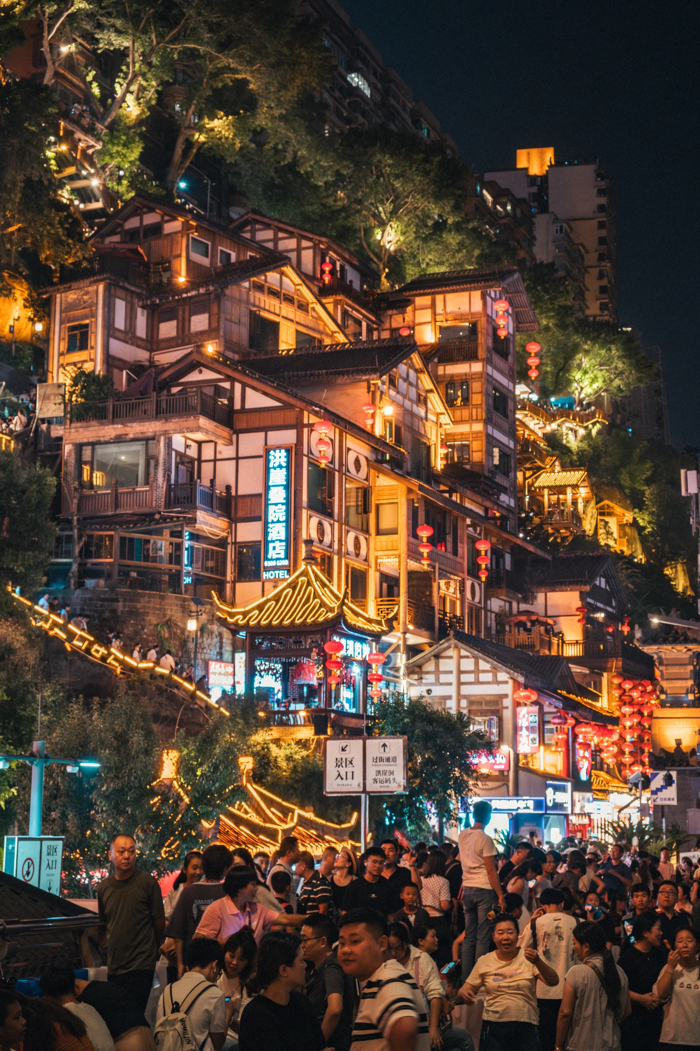 Hongya Cave district in Chongqing lit at night, tiered architecture clinging to a cliff, dense crowd at the foot of the site.