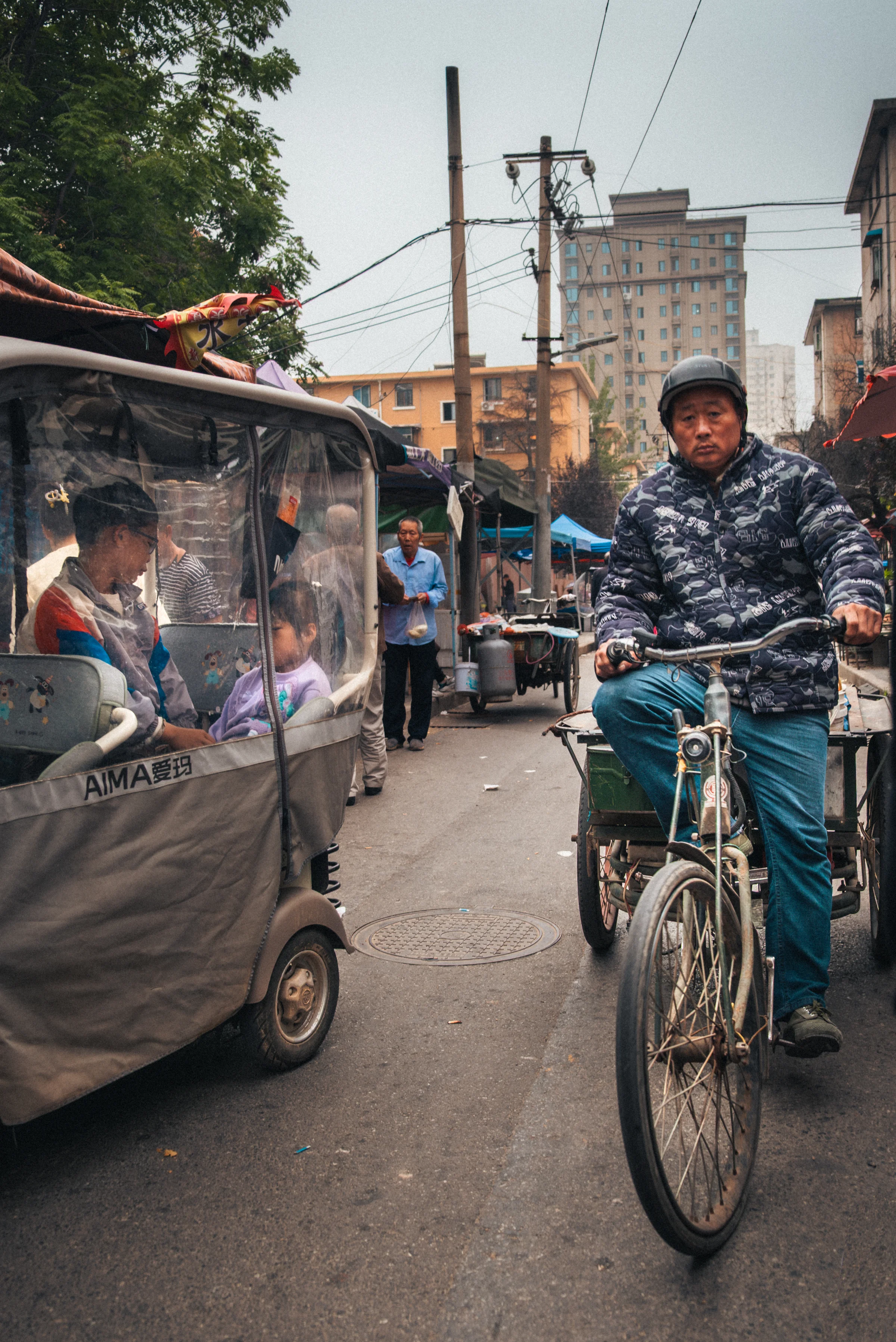 An alley in Xi'an on a gray morning, a man on a pedal tricycle on the right, an electric scooter carrying children behind a transparent tarp on the left.