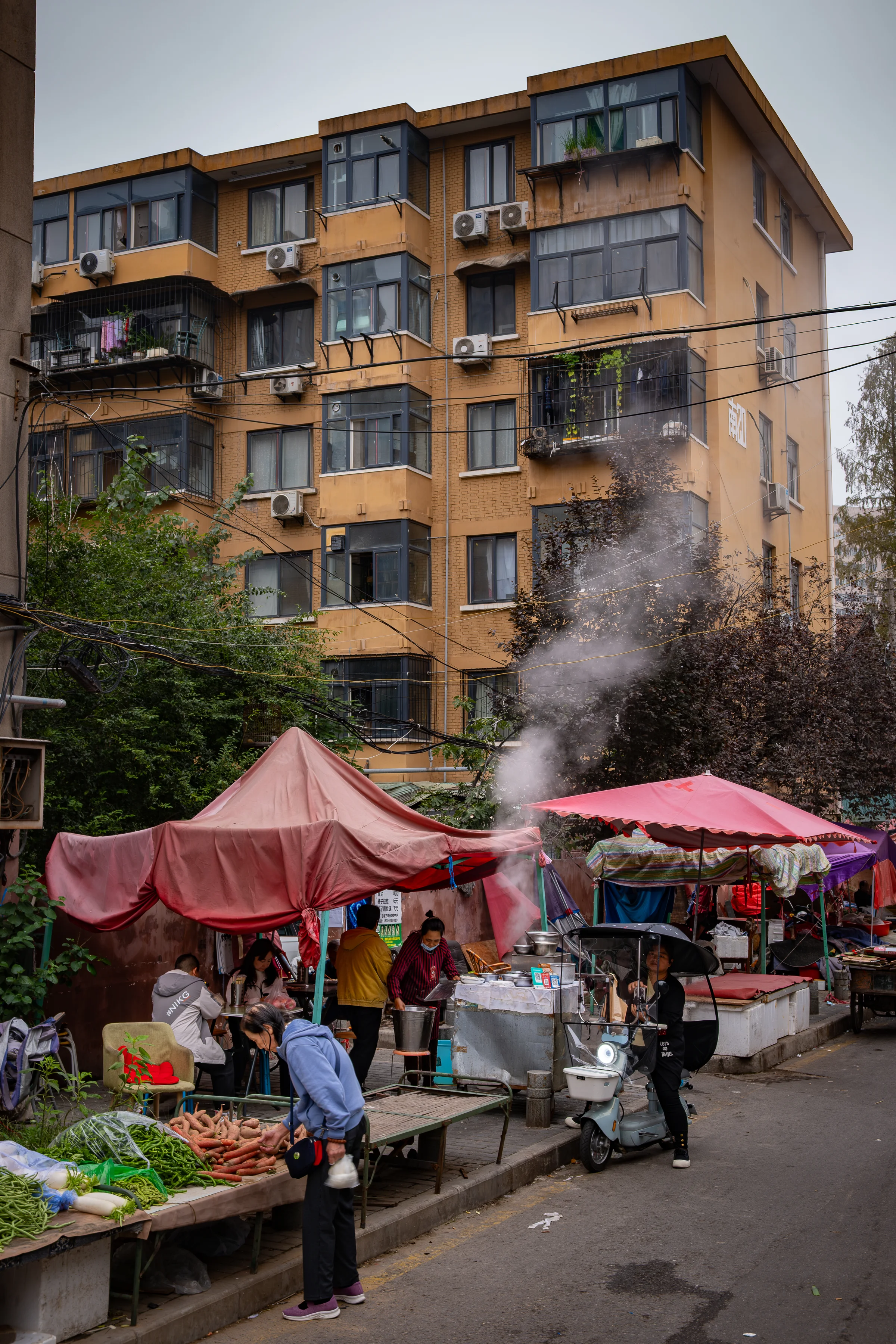 Morning market at the foot of a yellow residential building, pink tarps stretched above the stalls, steam from a cooking pot rising toward the electric wires.