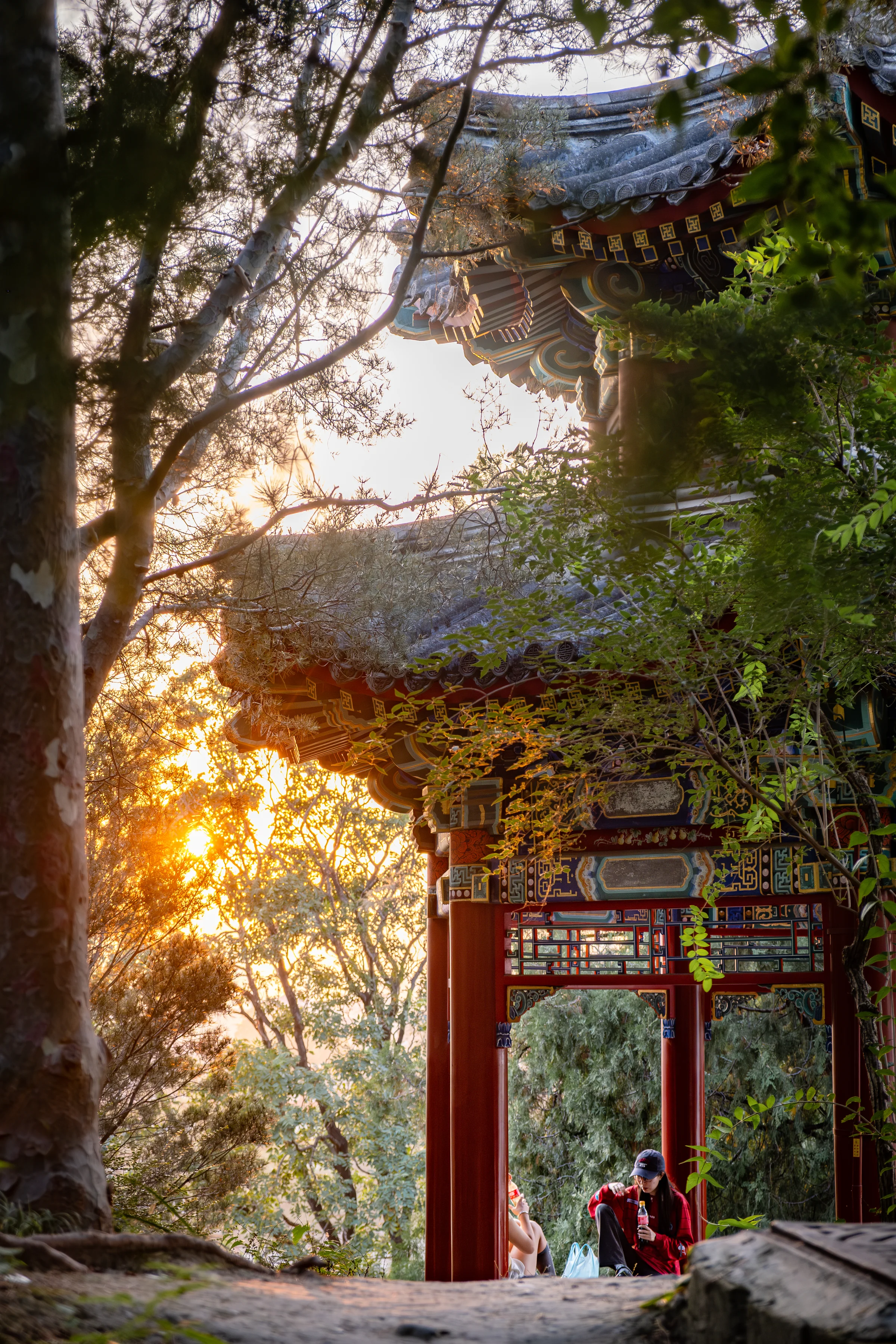 Imperial pavilion of Jingshan park bathed in golden sunset light, two young women sitting on the ground under the pavilion.
